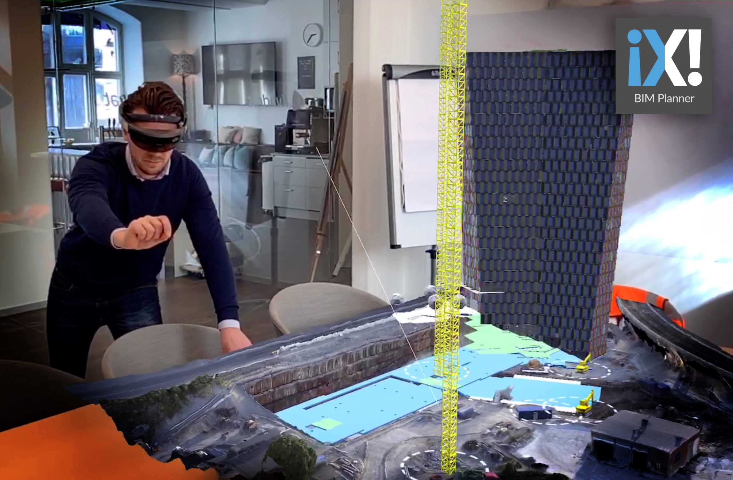 En man med HoloLens sträcker sig mot en holografisk platsmodell på ett konferensbord, med byggkran och etappindelning inlagda.