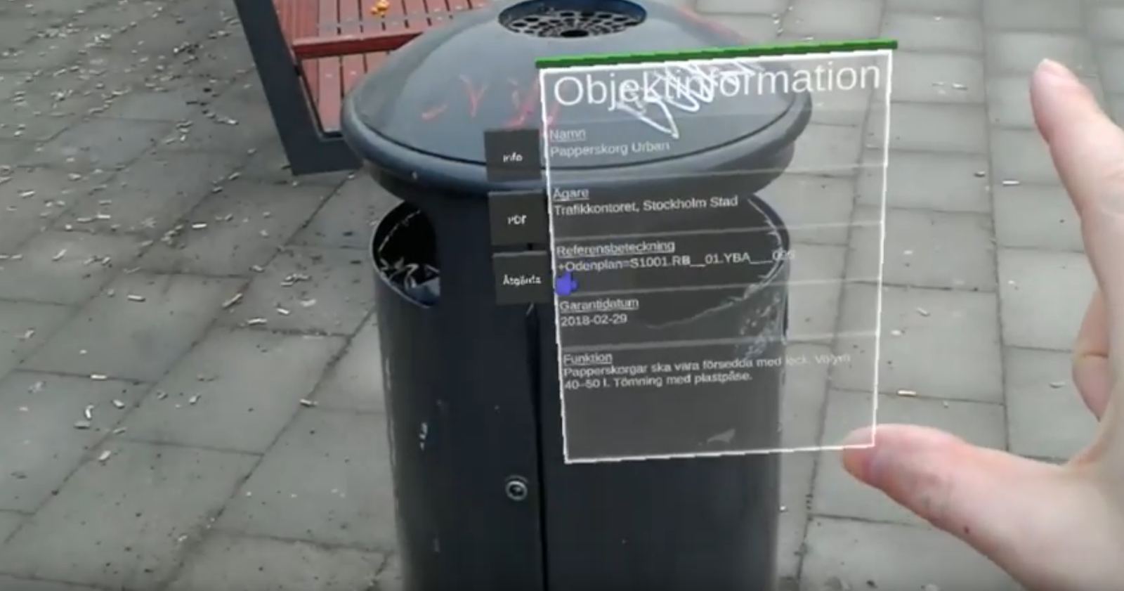 En papperskorg på en gata, med en informationsruta inlagd bredvid: Objektinformation, namn Papperskorg Urban, ägare Trafikkontoret Stockholms stad, referensbeteckning och garantidatum.