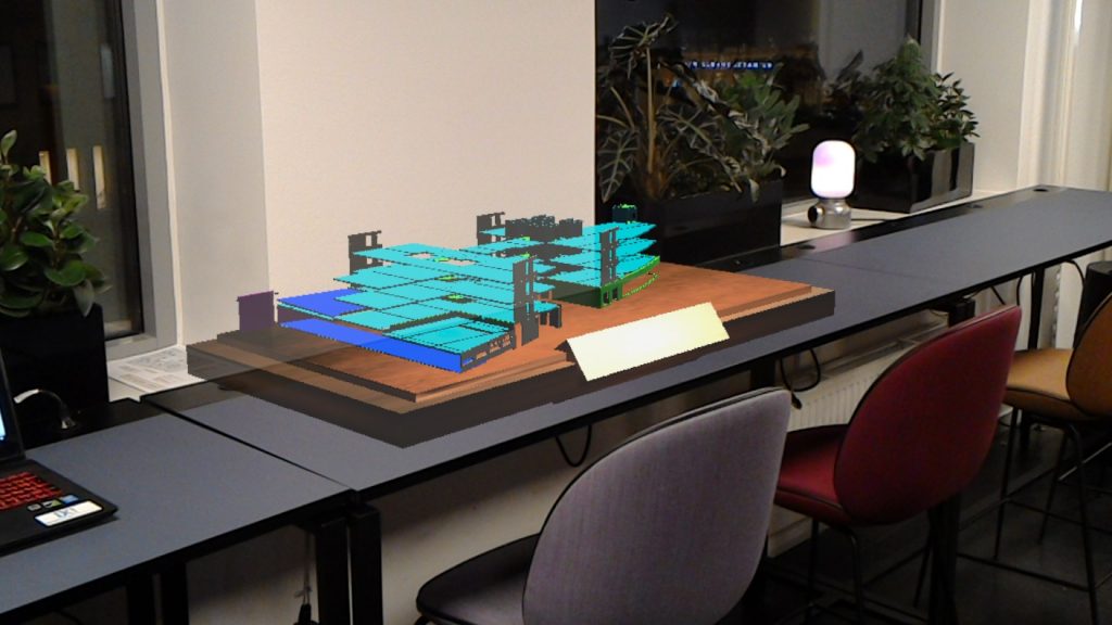 En byggnadsmodell i cyan och grönt ligger som hologram på ett kontorsbord bredvid en bärbar dator.