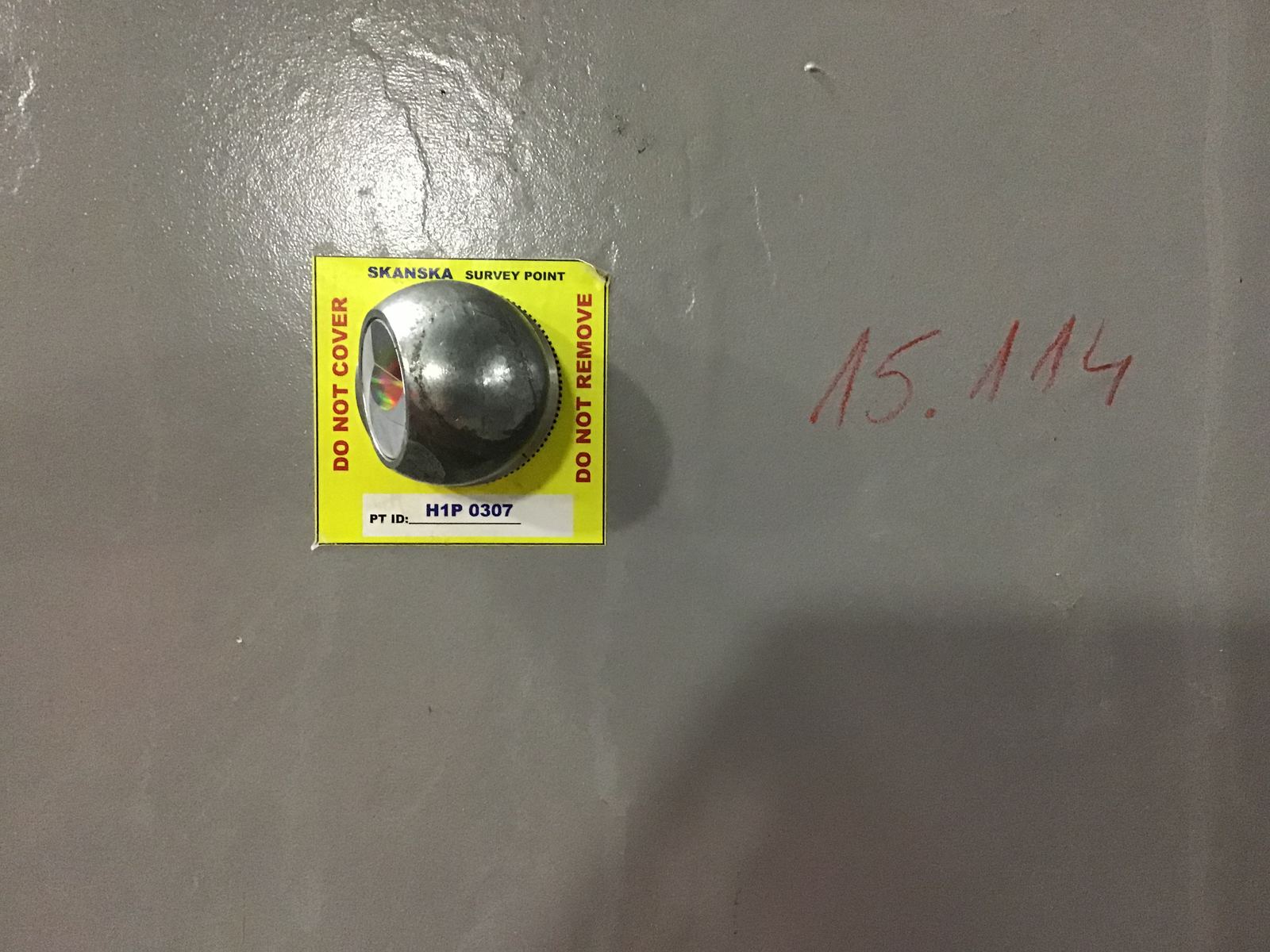 En metallhalvsfär monterad på en gul Skanska-etikett med texten SURVEY POINT, DO NOT COVER, DO NOT REMOVE och ett punkt-id, fäst på en betongvägg.