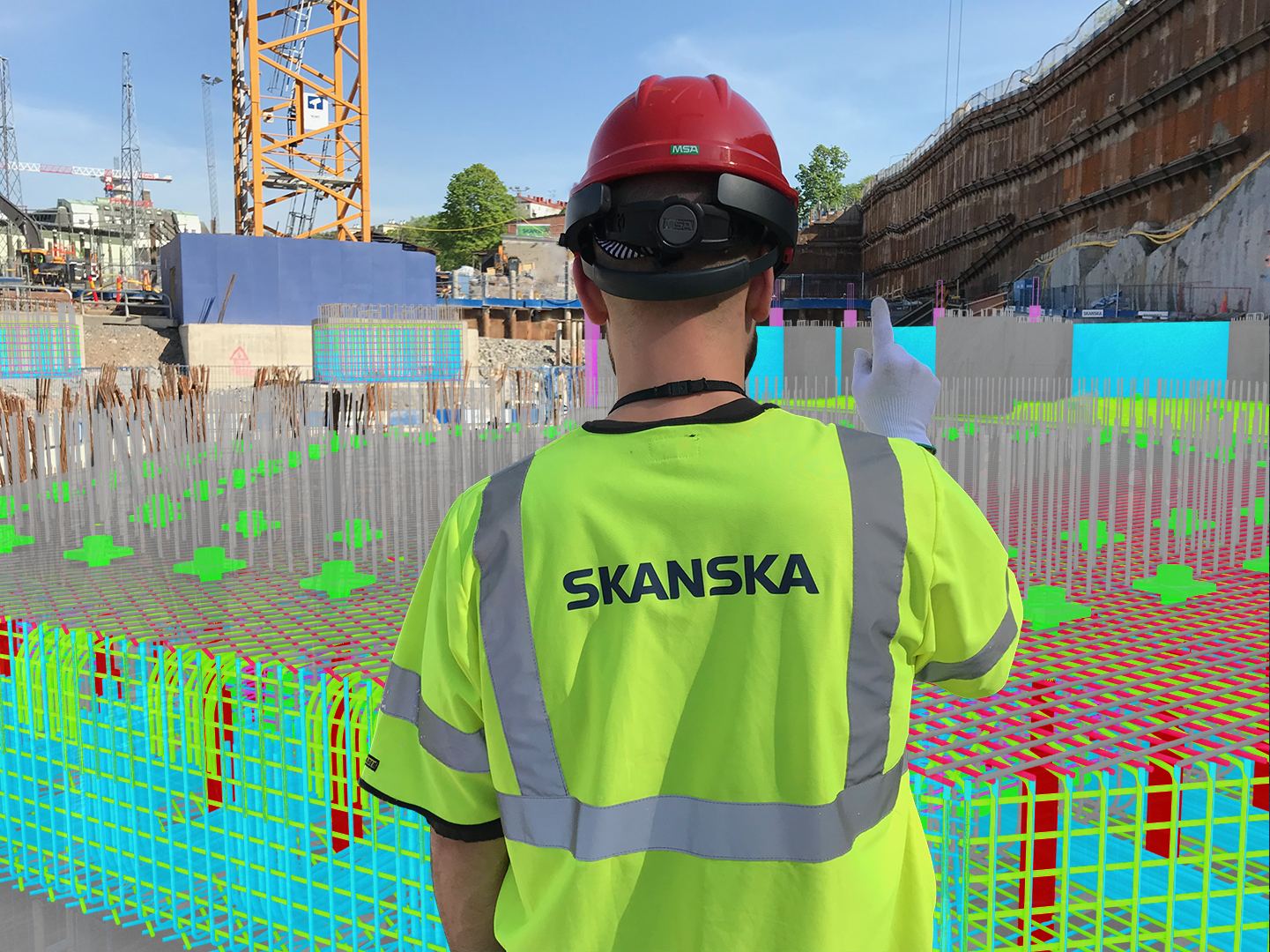 En byggnadsarbetare i Skanska-väst och röd hjälm med monterad HoloLens står med ryggen mot kameran vid en grundläggning. Över den verkliga armeringen ligger en modell i cyan, magenta och grönt.