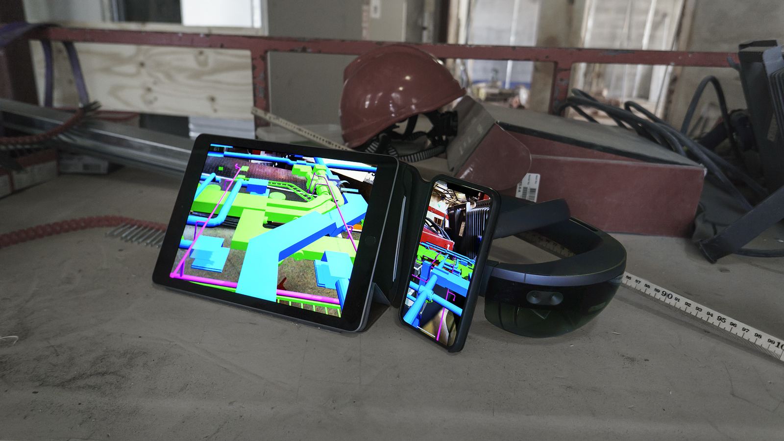 En läsplatta, en telefon och ett par HoloLens ligger på ett betonggolv. Alla tre visar samma installationsmodell i blått, grönt och magenta.
