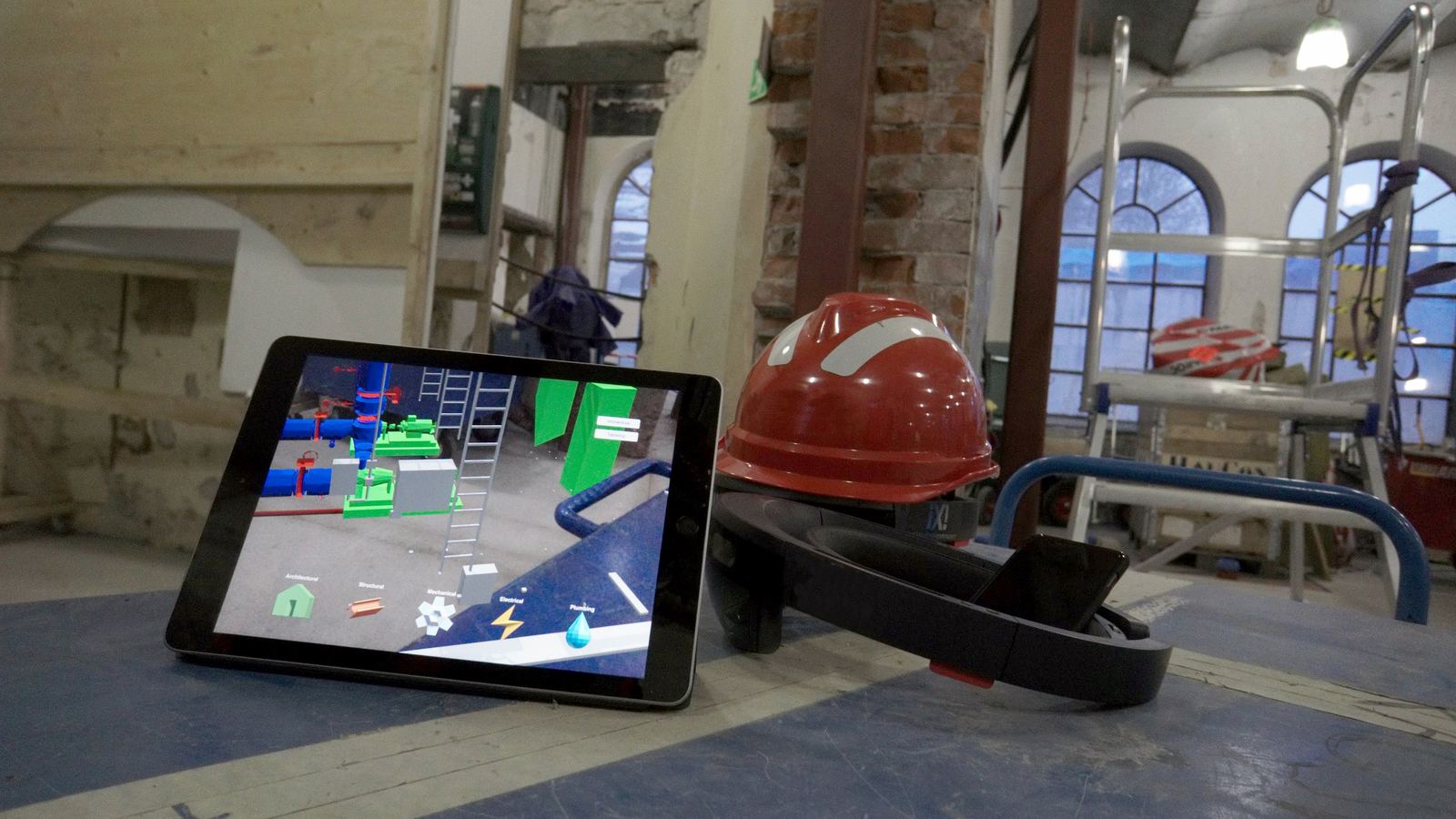 En iPad står lutad på ett golv i en äldre industribyggnad under ombyggnad, bredvid en hjälm och ett par HoloLens.